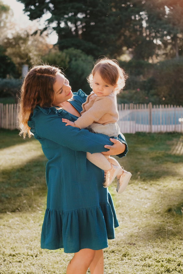 Maman qui porte son bébé dans un jardin, avec sa robe d'allaitement couleur bleu canard mi longue 